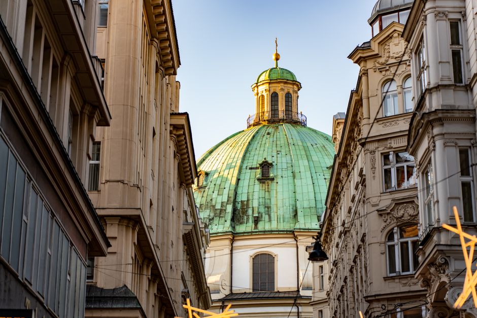 A detailed view of a historic cityscape featuring a prominent large green-domed building with gold and black decorative elements at the top, situated between classic European-style white stone buildin